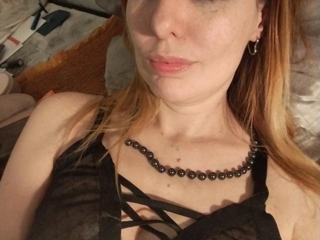 JujuBauva with pale skin and red hair, wearing a black beaded necklace and sheer black lingerie, showing her tits and pouty pink lips in a selfie.