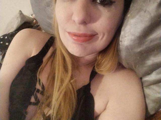 JujuBauva with blonde hair, wearing black lace lingerie, showing her tits, lying on a pillow, smiling with glossy pink lips, and looking at the camera.