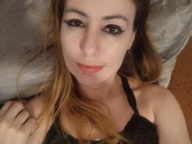 JujuBauva with light skin, brown eyes, and blonde hair, lying on a pillow, wearing black lingerie, smiling with glossy lips, and having a cute, sexy face.