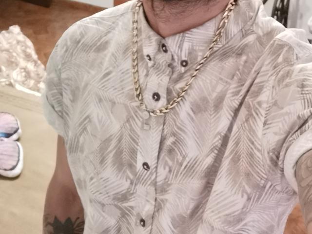 ZousGreen trägt ein gemustertes Hemd mit einer goldenen Kette, zeigt ein Tattoo auf seinem rechten Arm und im Hintergrund sind rosa Hausschuhe zu sehen..