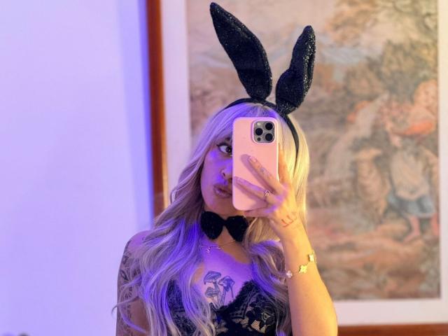 LunaDesire with blonde hair, black glitter bunny ears, black bowtie, lace top, holding phone, taking mirror selfie, purple light, tattoo on chest, gold bracelet.