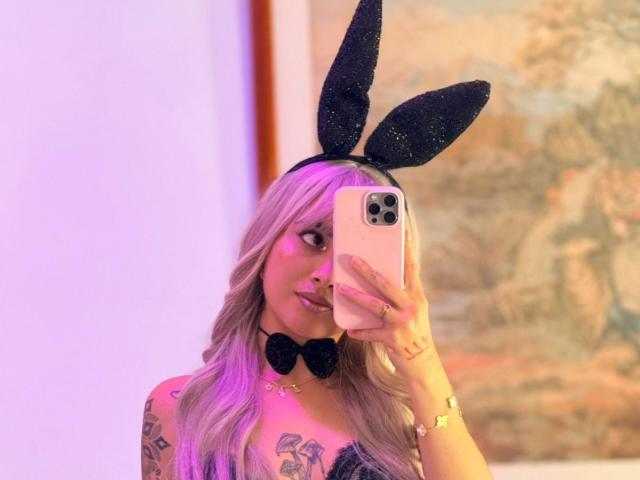 LunaDesire with pink hair, black bunny ears, bowtie, and tattoos, holding a pink phone, taking a mirror selfie, showing her tits and a subtle smile.