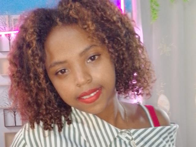 Nerina has curly brown hair, red lipstick, and is wearing a green and white striped shirt with a red strap. She's smiling softly with a cute, attractive face.