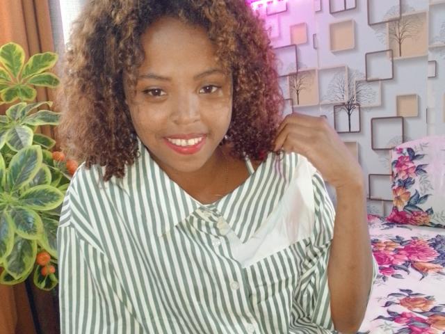 Nerina with curly brown hair, smiling, wearing a green-striped shirt, holding her shirt collar, standing by a plant, floral bedsheet, and wall with square patterns.