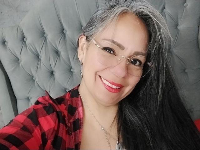 Laila Castillo, met grijs en zwart haar, draagt een bril, een rood-zwarte geruite blouse, glimlacht, heeft rode lippenstift op en een zilveren ketting, met op de achtergrond een gestoffeerd grijs hoofdeinde..