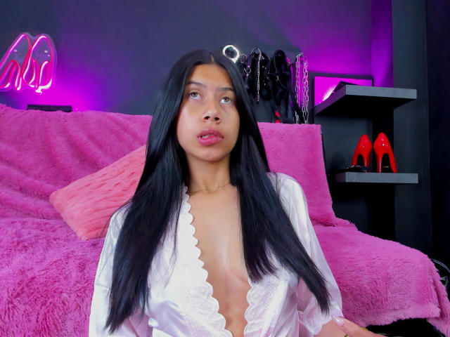 ChloeStone with long black hair, wearing a white lace robe, exposing her small tits, sitting on a pink bed, with purple lighting and black shelves with red heels.