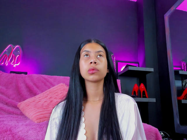 ChloeStone with long black hair, pouty lips, and a white robe, showing some cleavage. Neon pink and purple lights in the background. Red high heels on a shelf.