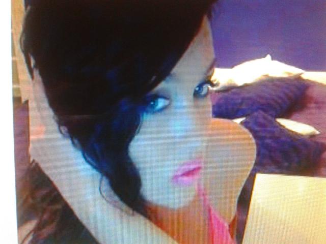 SexyLaylani-hot with dark wavy hair, blue eyes, pink lipstick, and a pink top, looking up with hands in her hair, sitting near a white box.