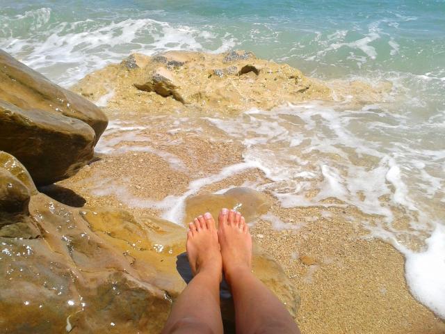 SexyLaylani-hot's bare feet on rocky beach, toes touching water, legs slightly apart, wet sand, small waves, ocean blue, rocks to the left, sunlight, clear sky, relaxed vibe.