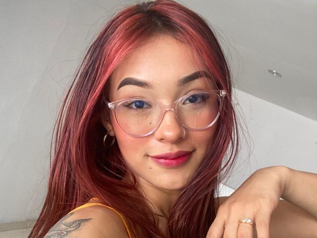 SashaHans has red hair, blue eyes, clear glasses, and pink lipstick. She's smiling with a small beauty mark on her right cheek and a tattoo on her left arm.