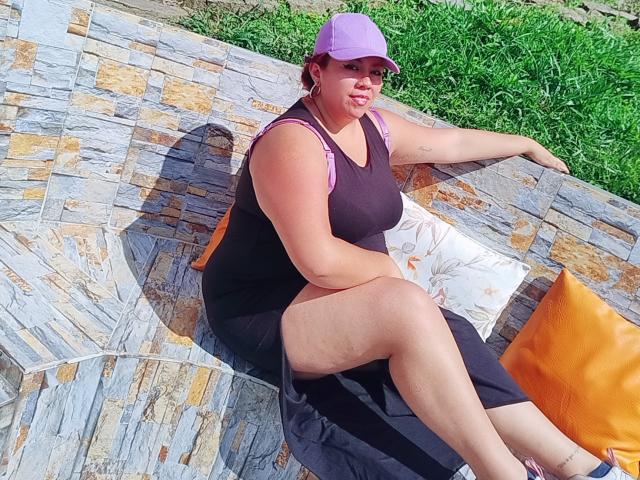 LexyDom is a chubby woman with big tits, wearing a purple cap, black dress, and pink tank top, sitting on a stone bench with orange and white pillows.