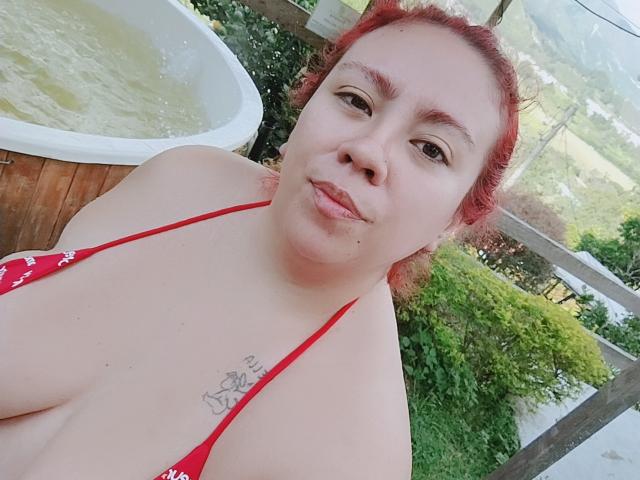 LexyDom with red hair, puffy lips, and a red bikini top showing her tits and a small tattoo on her chest, standing near a hot tub in a backyard.