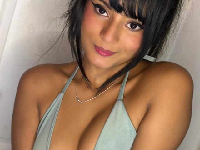 Marbela with dark hair, bangs, and pink lipstick, wearing a green halter top, showing her boobs, smiling, and looking at the camera, with a silver necklace.