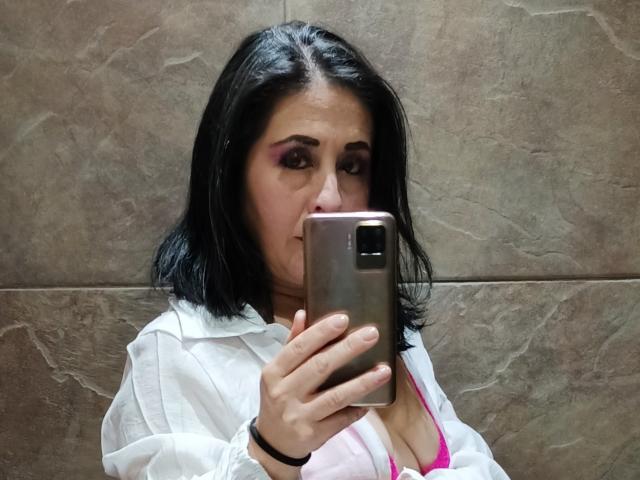 ScarletMilfX mit schwarzen Haaren, pinkem Augen-Make-up, einem weißen Hemd und einem pinken BH, hält ein Smartphone, macht ein Selfie und zeigt ihre Brüste, vor einer braunen, gefliesten Wand..