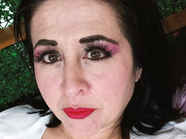 Close-up of ScarletMilfX with fair skin and dark hair. She has brown eyes, thick black eyebrows, and wears bright pink and purple eyeshadow with long, thick black eyelashes. Her red lipstick contrasts with her pale skin. The texture of her skin is visible with small bumps and blemishes. She's looking slightly to the right. Background includes green, textured pattern. Her expression is neutral. The image is focused on her face, with her white shirt in the bottom left corner.