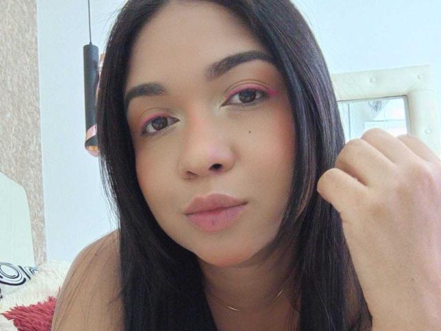 AliceSmity has medium brown skin, black straight hair, pink eyeshadow, and a small beauty mark near her nose. She's making a slight pout with her lips, touching her hair.