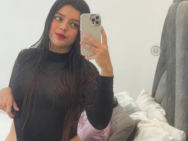 Violetta Rouse, con lunghi capelli neri, indossa una maglia nera trasparente, tiene in mano un telefono con una custodia scintillante, mostra il seno, ha il rossetto rosso e un sorriso sicuro, in un selfie allo specchio..