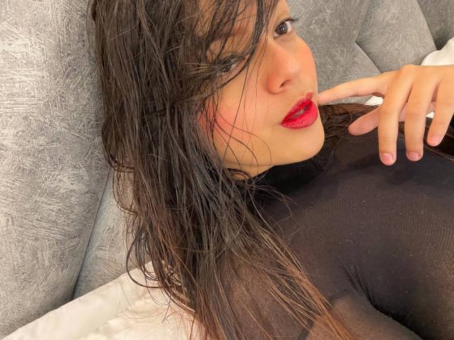 Violetta Rouse hat feuchtes, braunes Haar, roten Lippenstift und ein schwarzes, durchsichtiges Oberteil, wobei ihre Finger in der Nähe ihres Mundes sind. Sie liegt auf einem grauen, strukturierten Sofa. Ihr Hintern ist leicht sichtbar..