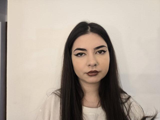 KiyraMoon heeft lang zwart haar, donkere wenkbrauwen, eyeliner met een vleugje, bruine lippenstift en een neusring. Ze draagt een witte blouse en kijkt recht in de camera..