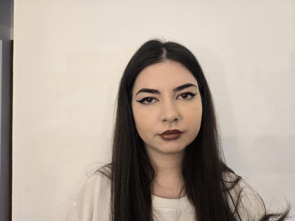 KiyraMoon heeft lang zwart haar, donkere wenkbrauwen, eyeliner met een vleugje, bruine lippenstift en een neusring. Ze draagt een witte blouse en kijkt recht in de camera..