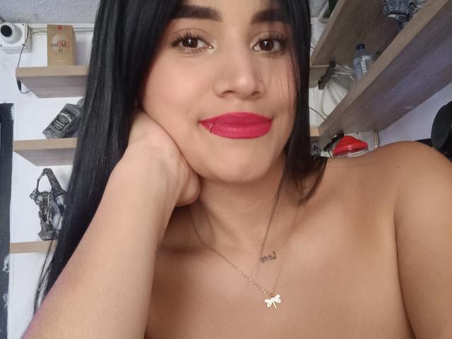 Alexandra Fode, oben ohne, mit langen schwarzen Haaren, braunen Augen, rotem Lippenstift, einem Nasenring und einer Schmetterlingskette, lehnt ihren Kopf in ihre Hand und lächelt. Brüste sind sichtbar..