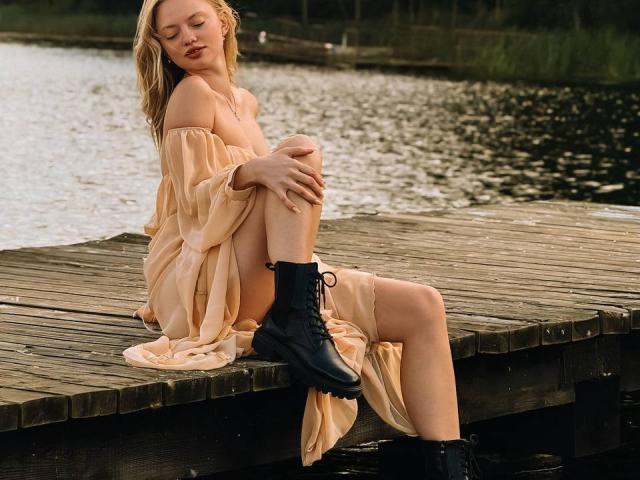 CrystalHope with blonde hair, off-shoulder beige dress, and black boots, sitting on wooden dock, legs crossed, eyes closed, looking relaxed, her bare tits slightly visible under dress.