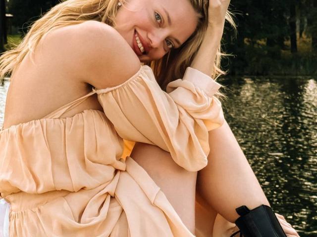 CrystalHope with blonde hair, smiling, wearing a peach off-shoulder dress, sitting by a lake, knees up, hand in hair, black boots, showing her bare shoulders and upper thighs.