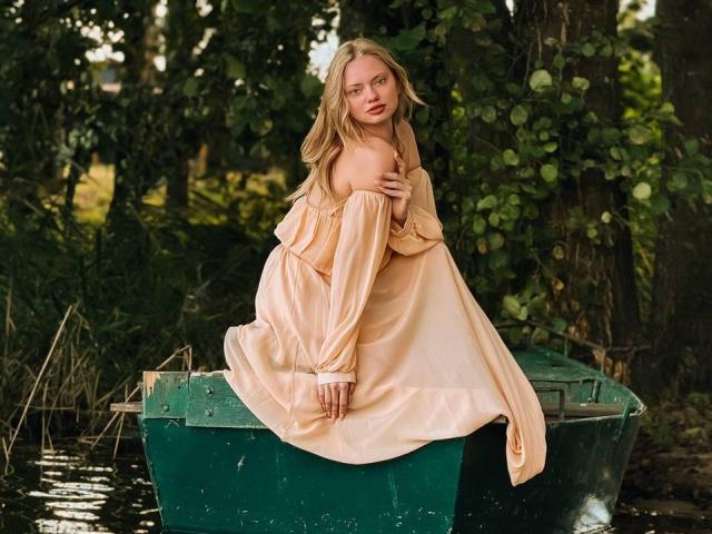 CrystalHope with blonde hair, wearing a pale pink off-shoulder dress, sits on a green boat, hands on chest, looking back with a slight smile, in a forest setting.
