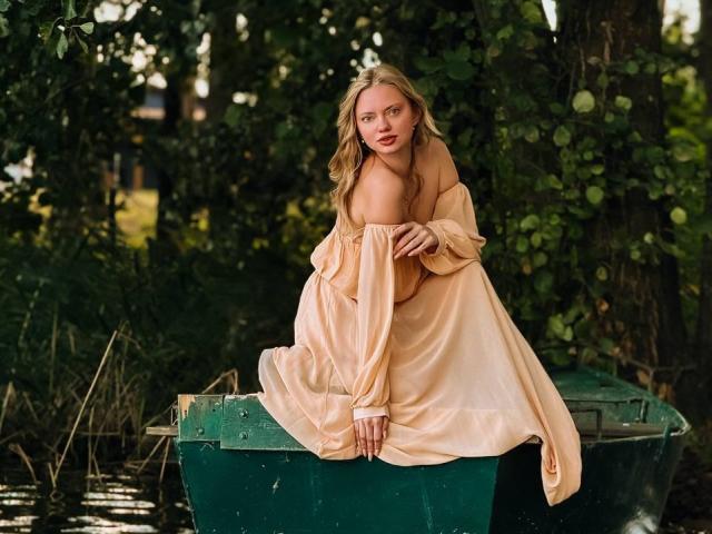 CrystalHope with blonde hair, red lips, and pale skin, sitting on a green boat in a peach off-shoulder dress, looking slightly to the side with a serious expression.