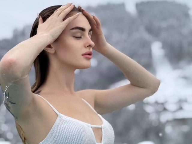 PlayfulfBoo with hands in brown hair, wearing white mesh top, showing small tits, tattoo on left arm, pale skin, pink lips, and closed eyes in foggy, rocky landscape.
