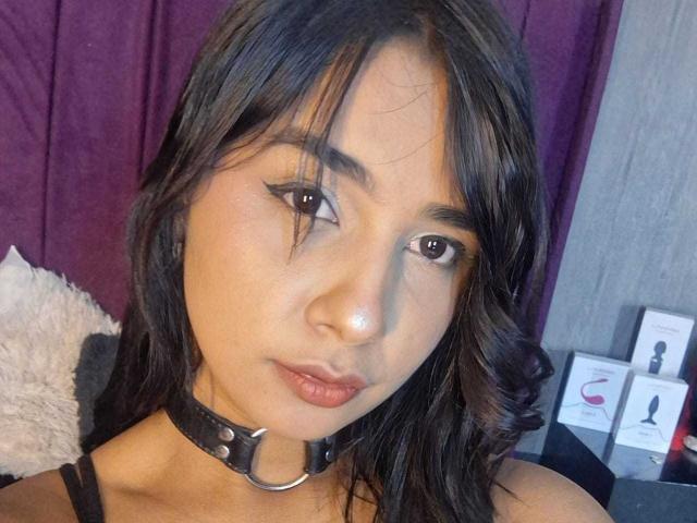 MariamLara has dark wavy hair, brown eyes, and wears a black choker with a ring. She has a subtle nose sparkle and slightly parted lips. Boobs are not visible.
