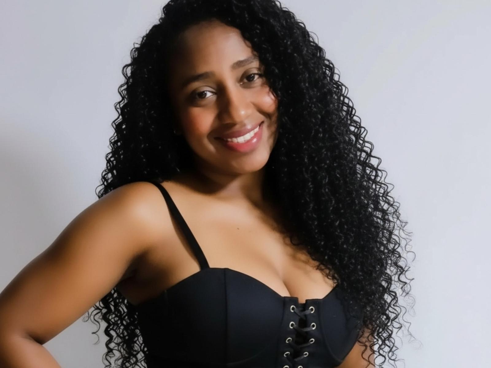 BlackPussySweet with long curly black hair, smiling, wearing a black bra with lace-up front, showing her tits, standing against a plain white background.
