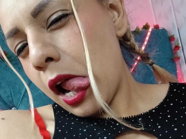 KathaleyaGrayyAss with eyes closed, tongue out, and red lipstick, wearing black polka-dot top, blonde braids, and red bra strap, looking slightly annoyed.