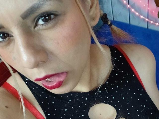 KathaleyaGrayyAss with blonde hair, red lipstick, and black polka-dot top, has her tongue out, showing her red bra straps and a cut-out neckline, looking at the camera.