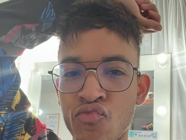 MiloJhones with glasses, puffy lips, and a slight mustache, touching his messy brown hair, wearing a colorful shirt, in a well-lit mirror selfie.
