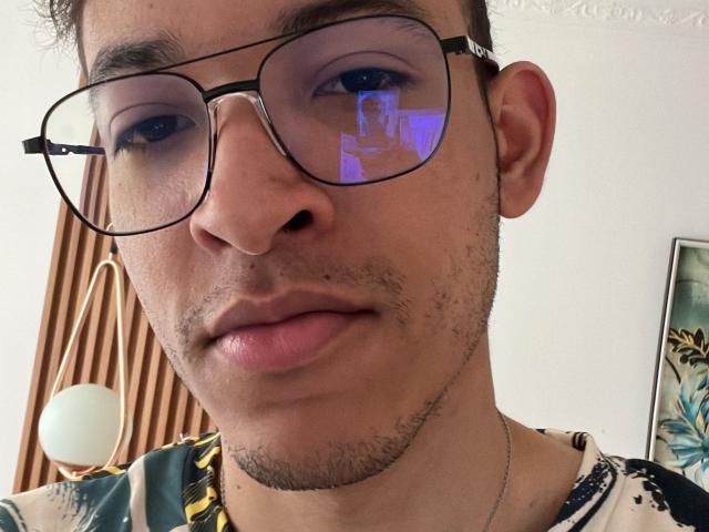 MiloJhones with glasses, slightly parted lips, and a reflection of a blue-lit room in the lenses. Short curly hair, light stubble, and a floral shirt. Casual, cute.