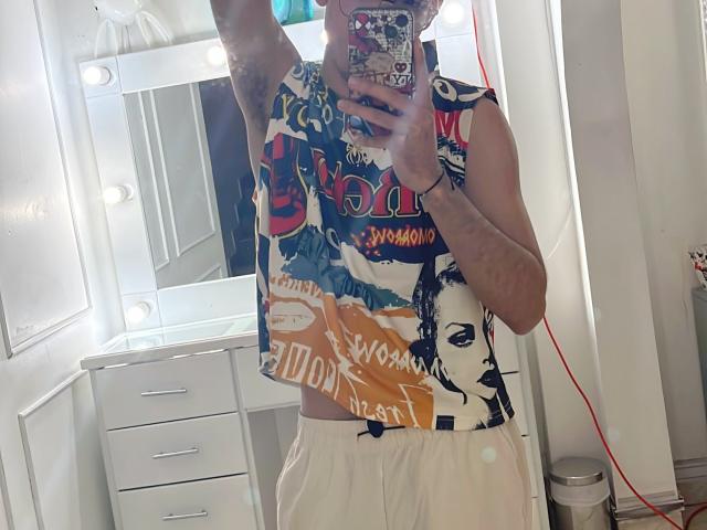 MiloJhones in a colorful sleeveless shirt with graphic prints, white pants, holding a phone in a mirror selfie, visible tattoos, mirror lights, and a red cord.