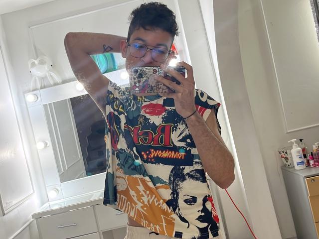 MiloJhones with glasses, short curly hair, tattoo on arm, wearing colorful graphic tee, holding phone in mirror selfie, lights in background, white bathroom, visible midriff, phone case with design.
