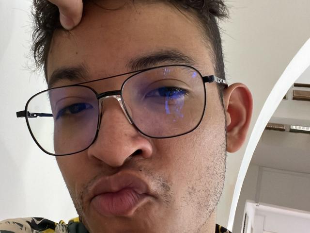 MiloJhones with puffy lips, wearing oversized glasses, has a hand in his curly hair, blue reflection in his eyes, slight stubble, and a neutral expression.