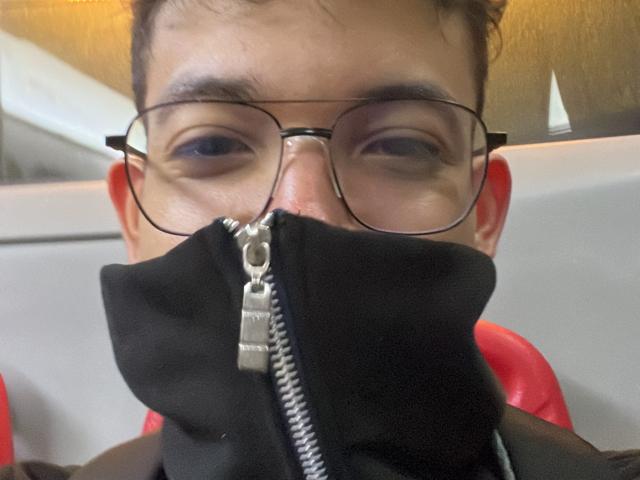 MiloJhones with glasses, wearing a black mask with a metal zipper, his nose and eyes visible, red chairs in the background, slightly messy brown hair.
