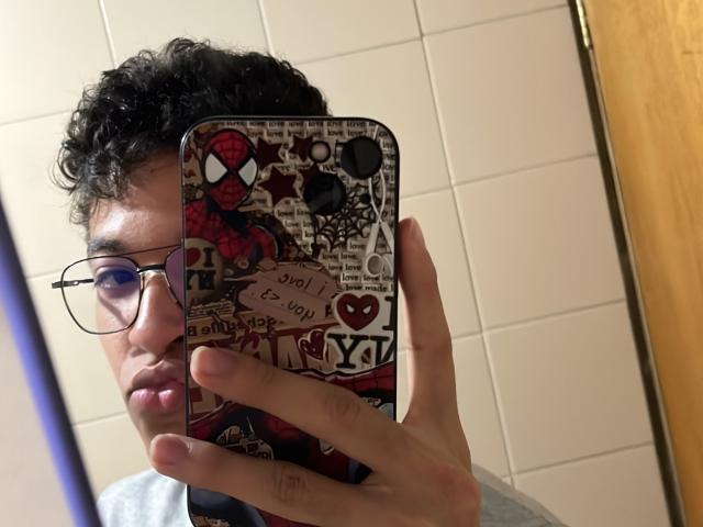 MiloJhones with curly black hair, glasses, holding a phone with a colorful, heart-themed case, taking a mirror selfie in a bathroom. Phone case has "I ♥ NY" text.