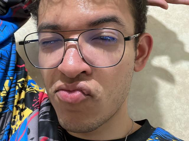 MiloJhones with shiny blue eyeshadow, puffy pink lips, and thick-rimmed glasses, wearing a colorful, patterned shirt. His nose is shiny, and he's sticking out his bottom lip.