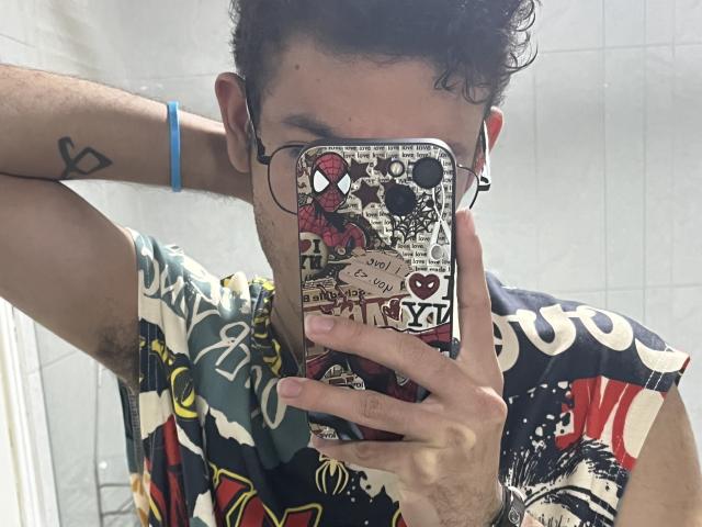 MiloJhones with curly brown hair, wearing a colorful, patterned shirt, holding a phone with Spider-Man and sticker-covered case, taking a mirror selfie, blue bracelet on right arm.