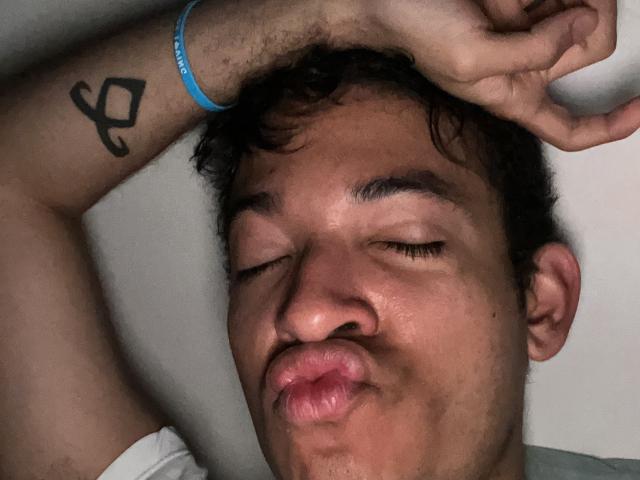 MiloJhones with his eyes closed, puffy lips in a kissing gesture, right arm raised, showing a black tattoo of an "A" on his wrist. Wearing a blue bracelet.