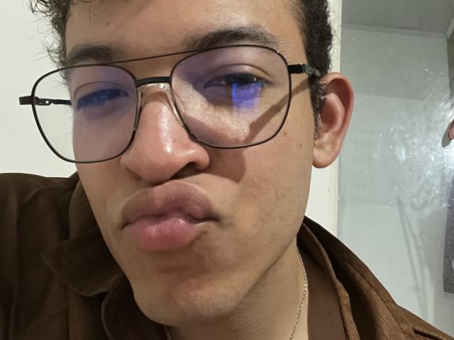 MiloJhones with blue-tinted glasses, puffy lips, and brown jacket, showing a slight pout. Short curly hair, light skin, and a small nose. Blue reflection in glasses.