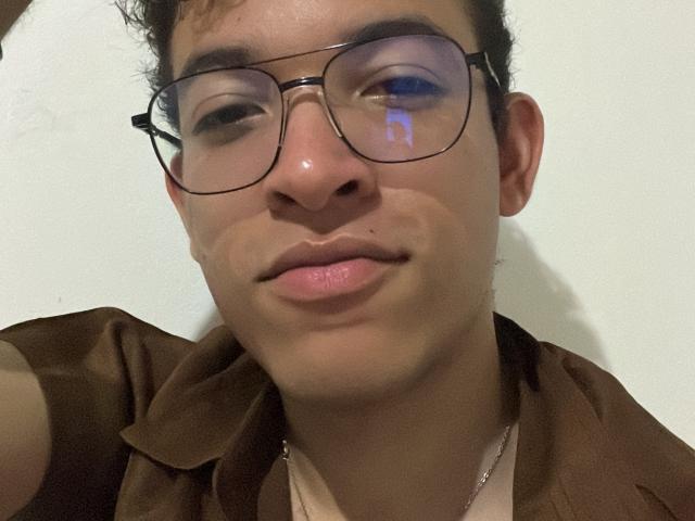 MiloJhones with glasses, brown shirt, and necklace, has a slight smile, a small nose piercing, and a light blue reflection in his right eye. His skin is light brown.