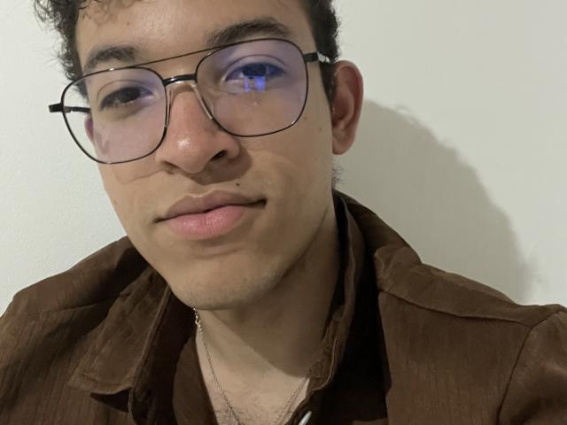 MiloJhones has curly brown hair, wears glasses, and a brown shirt, with a small mole on his right cheek and a subtle, relaxed smile. His ass is not visible.