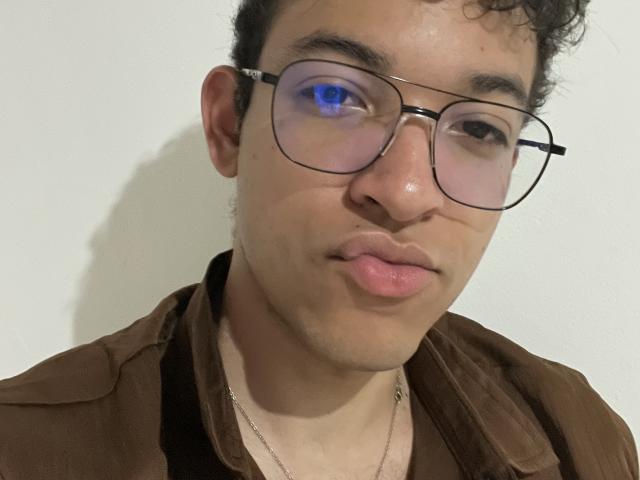 MiloJhones with curly brown hair, glasses, and puffy pink lips, wearing a brown shirt, showing a slightly goofy expression. His ass is not visible.