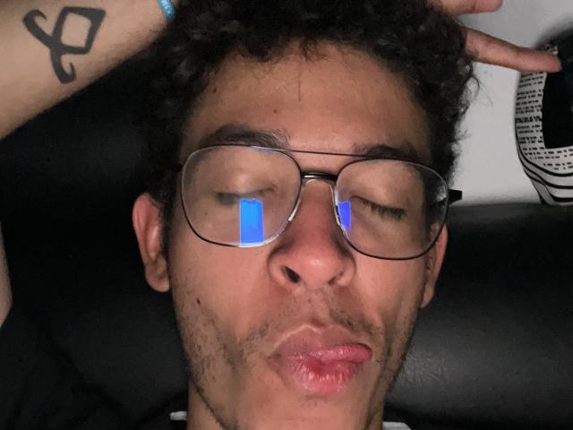 MiloJhones with curly hair, glasses reflecting a blue light, puckering lips, black tattoo on right arm, holding head with left hand, wearing black shirt, dark background.