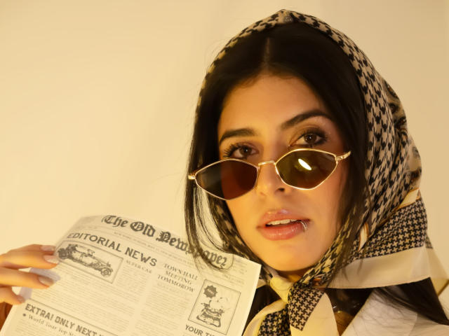 MiluSky with dark hair, wearing a patterned headscarf and aviator sunglasses, holding a newspaper, has a slightly open mouth with a small silver lip piercing.